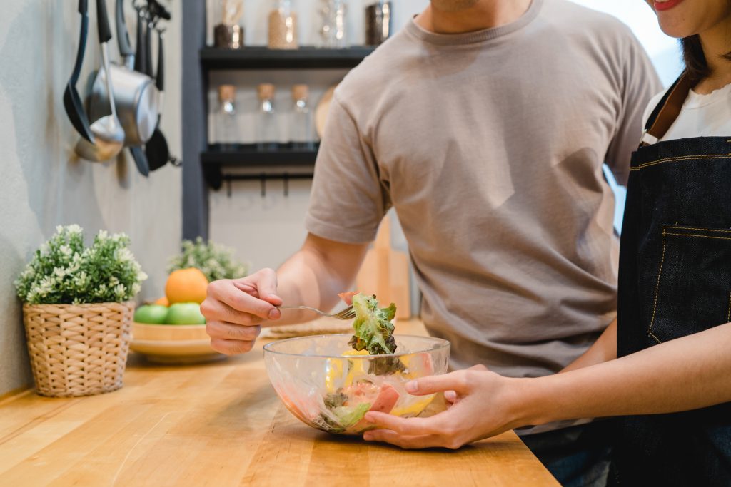 Pareja de hombre y mujer están de pie en la cocina preparando y comiendo una ensalada saludable. 